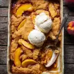 Golden brown peach cobbler with ice cream in a baking dish on rustic wooden table