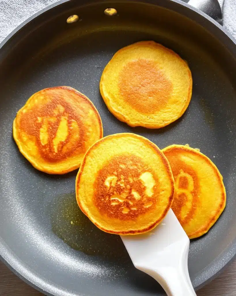 Fluffy pumpkin cottage cheese pancakes cooking in a non-stick skillet with one pancake lifted by a white spatula.