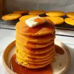 Tall stack of pumpkin cottage cheese pancakes topped with butter and maple syrup on a plate with extra pancakes in the background.