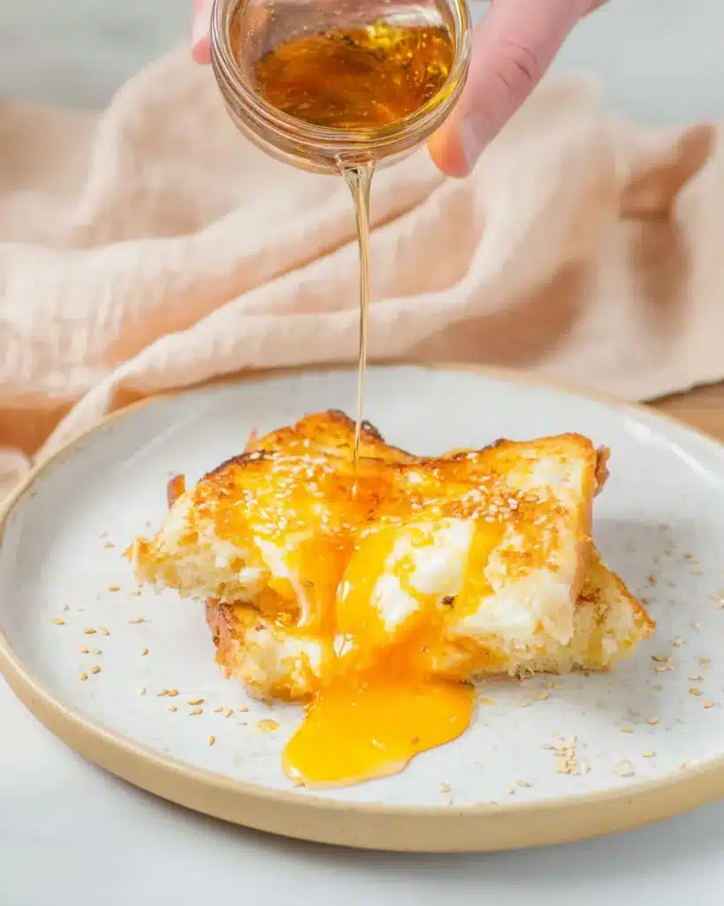 Pouring syrup over air fryer egg and cheese toast with runny yolk and sesame seeds on a white plate