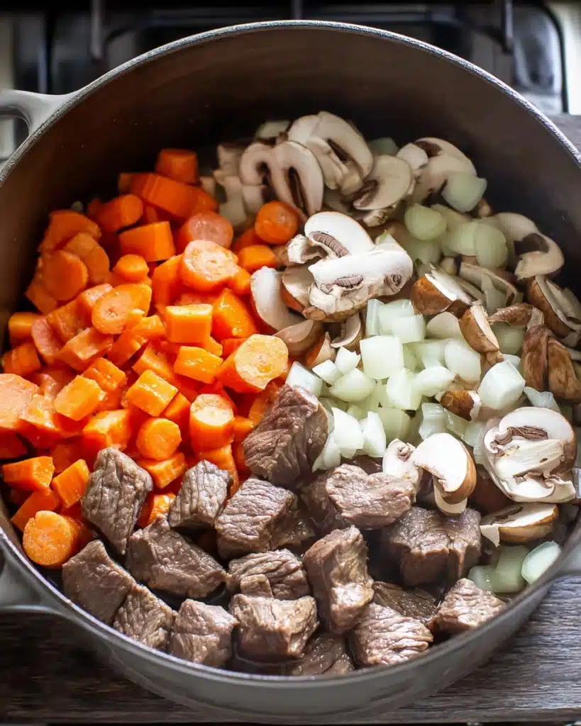 Beef barley soup ingredients with carrots, onions, and mushrooms in Dutch oven