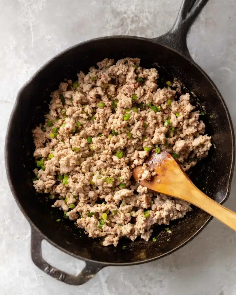 Ground pork browning in a cast iron skillet with wooden spoon