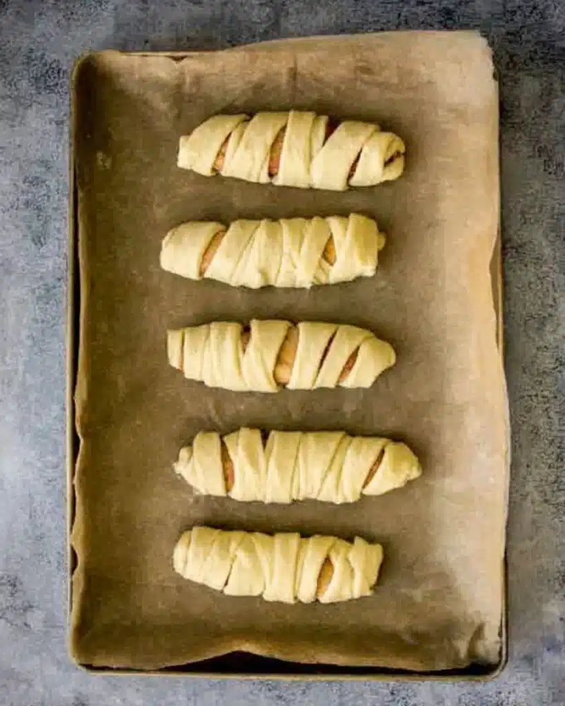 Uncooked Halloween sausage mummies wrapped in puff pastry on baking tray