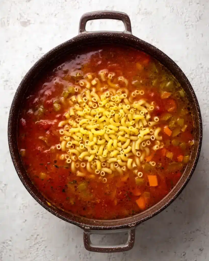 Uncooked pasta added to simmering tomato broth for minestrone soup