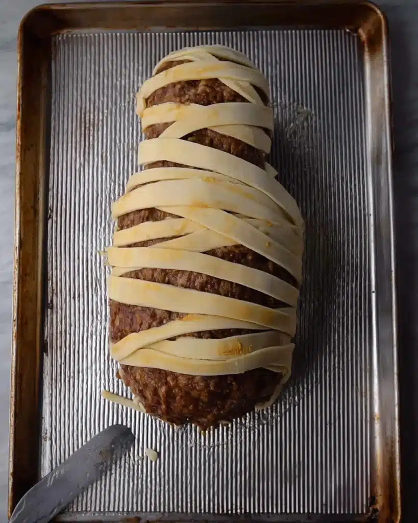 Partially wrapped mummy meatloaf with puff pastry strips on a baking sheet
