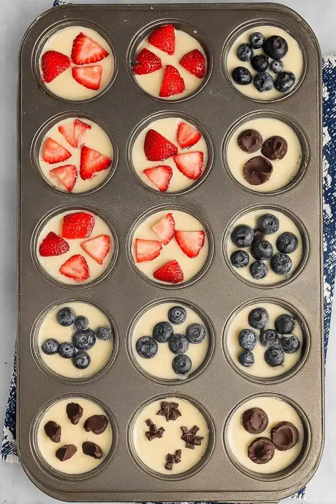 Raw pancake mini muffins in a tin, topped with strawberries, blueberries, and chocolate chips, ready for baking.