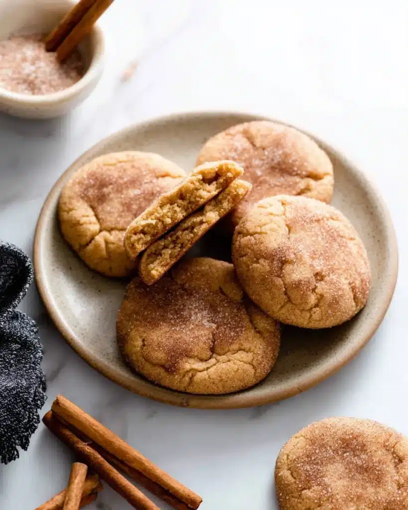 Plate of snickerdoodle cookies with one broken to reveal the soft, chewy interior, and cinnamon sticks.