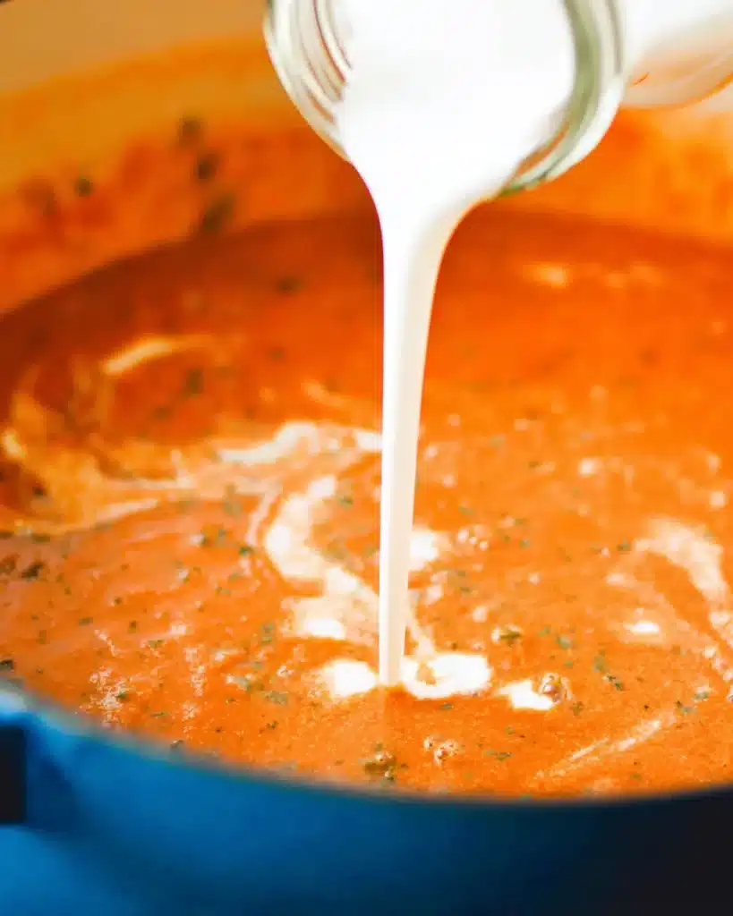 Pouring cream into tomato basil soup in a blue pot